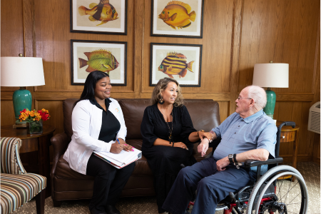 Allied health professional discussing care plan with older man and family member in a relaxed home setting.